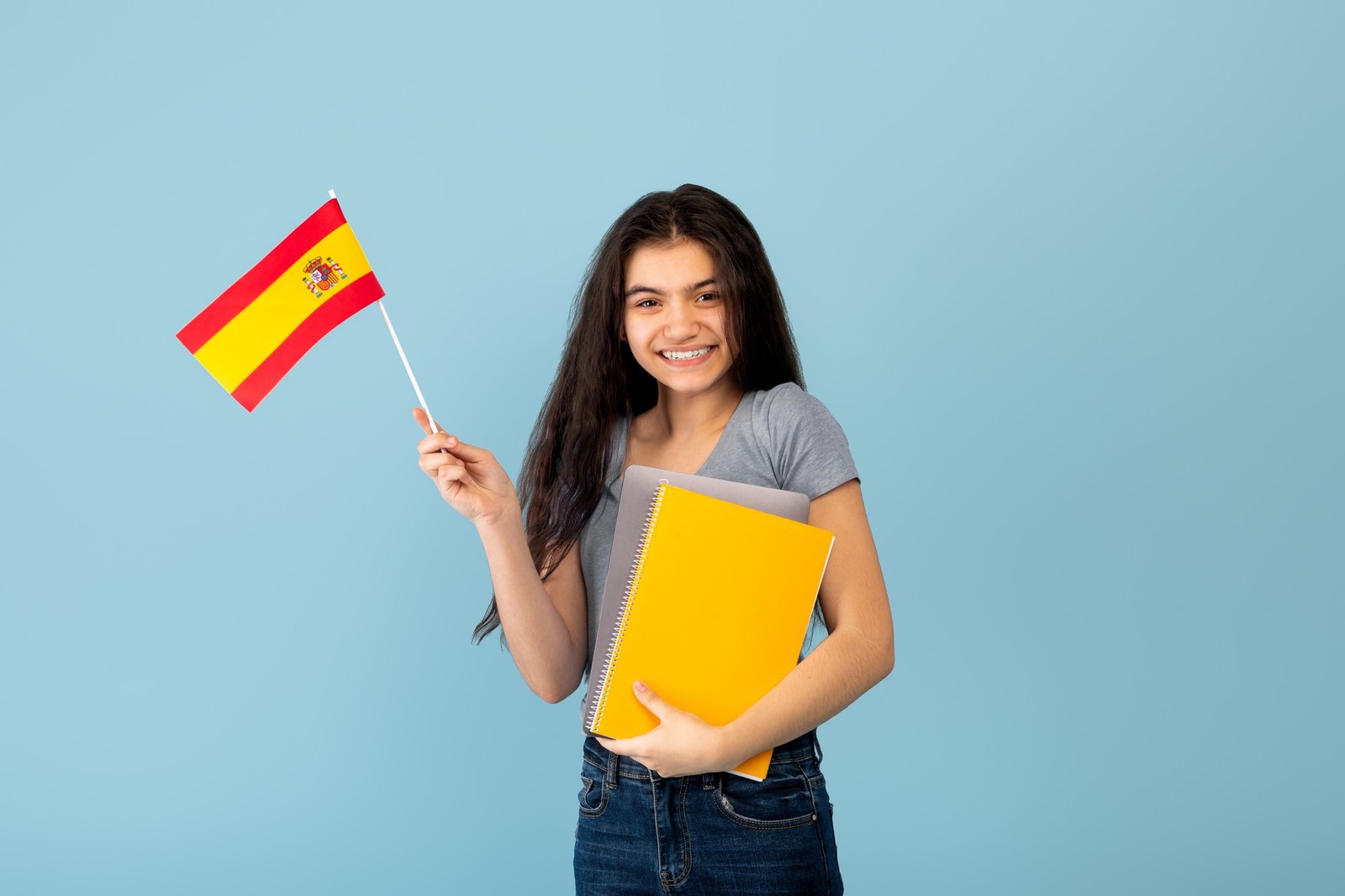 Cheerful Indian teen exchange student with notebook and laptop pc holding flag of Spain on blue