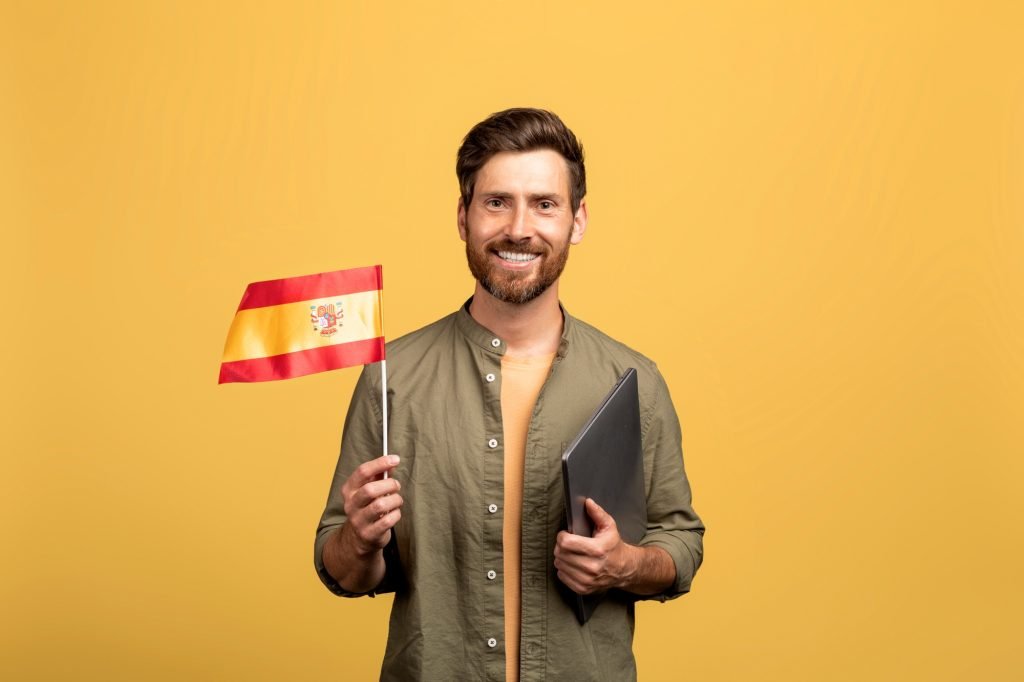 Cheerful middle aged man holding Spain flag and laptop, posing on yellow studio background and