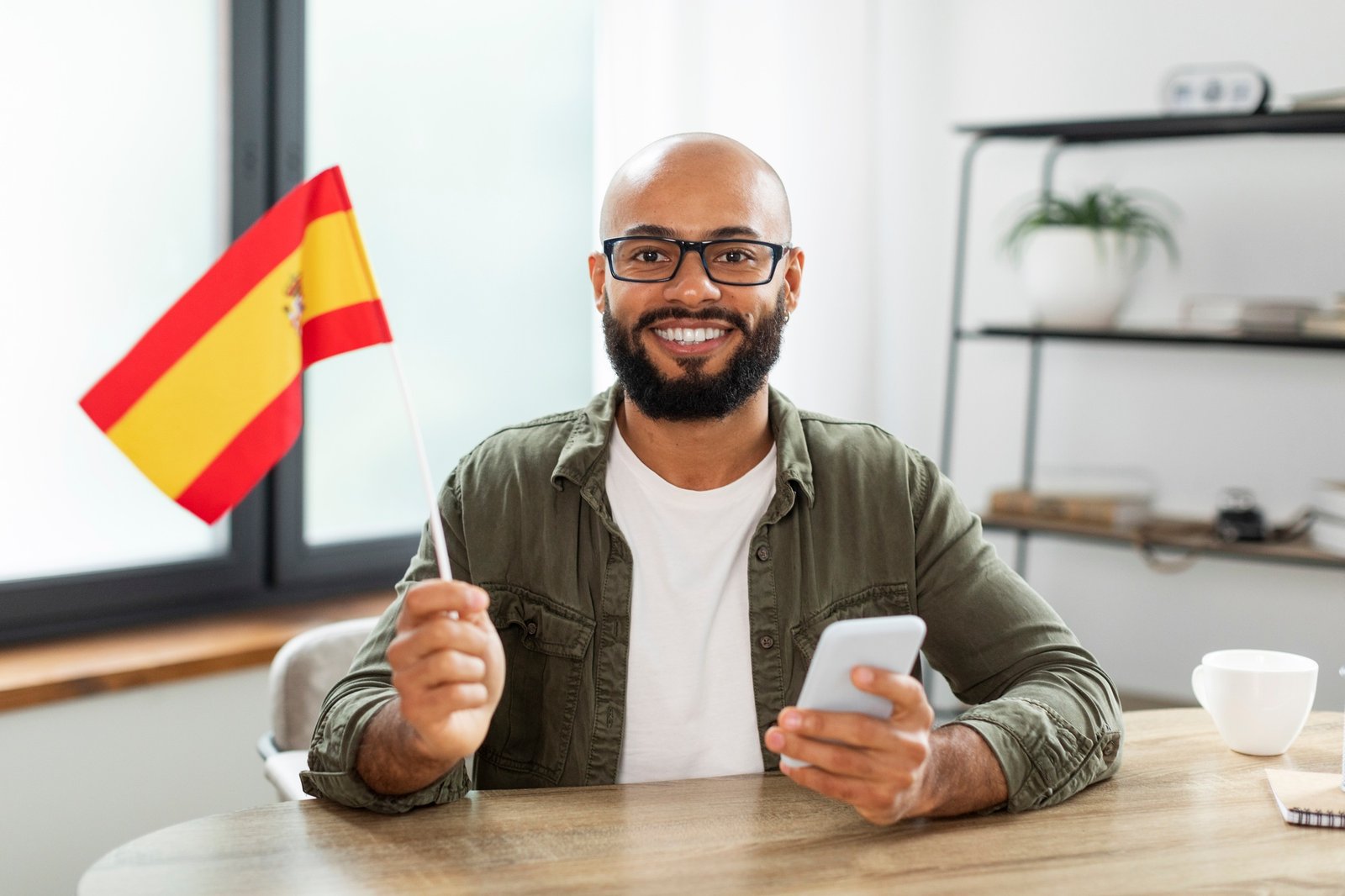 Excited male tutor sitting at table with flag of Spain and using smartphone, looking and smiling at