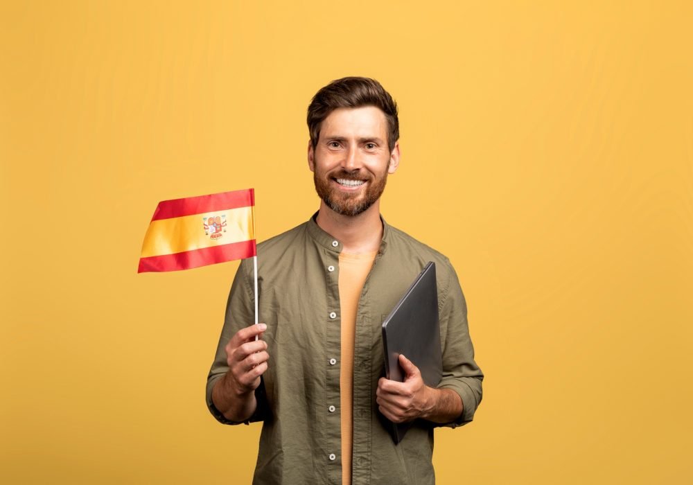 cheerful-middle-aged-man-holding-spain-flag-and-laptop-posing-on-yellow-studio-background-and.jpg