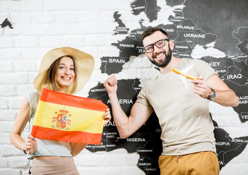 Couple of young travelers standing with spanish flag near the wall with world map, dreaming about summer vacations in Spain